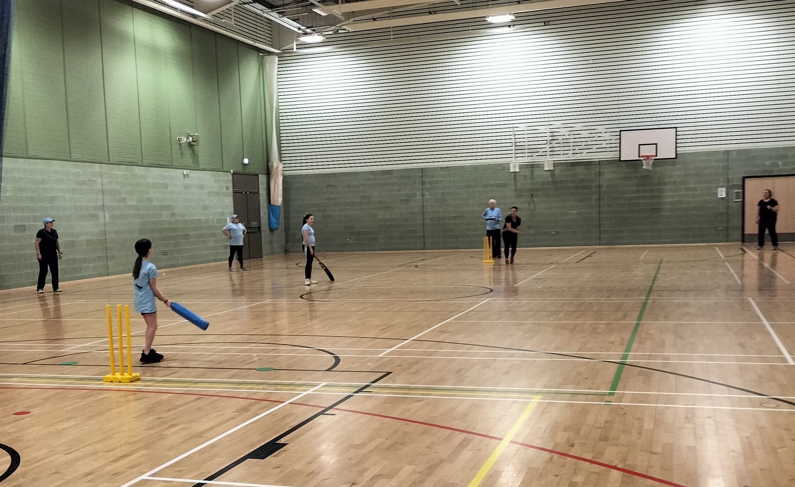 People playing cricket in large hall that has a wooden floor and light green walls, 