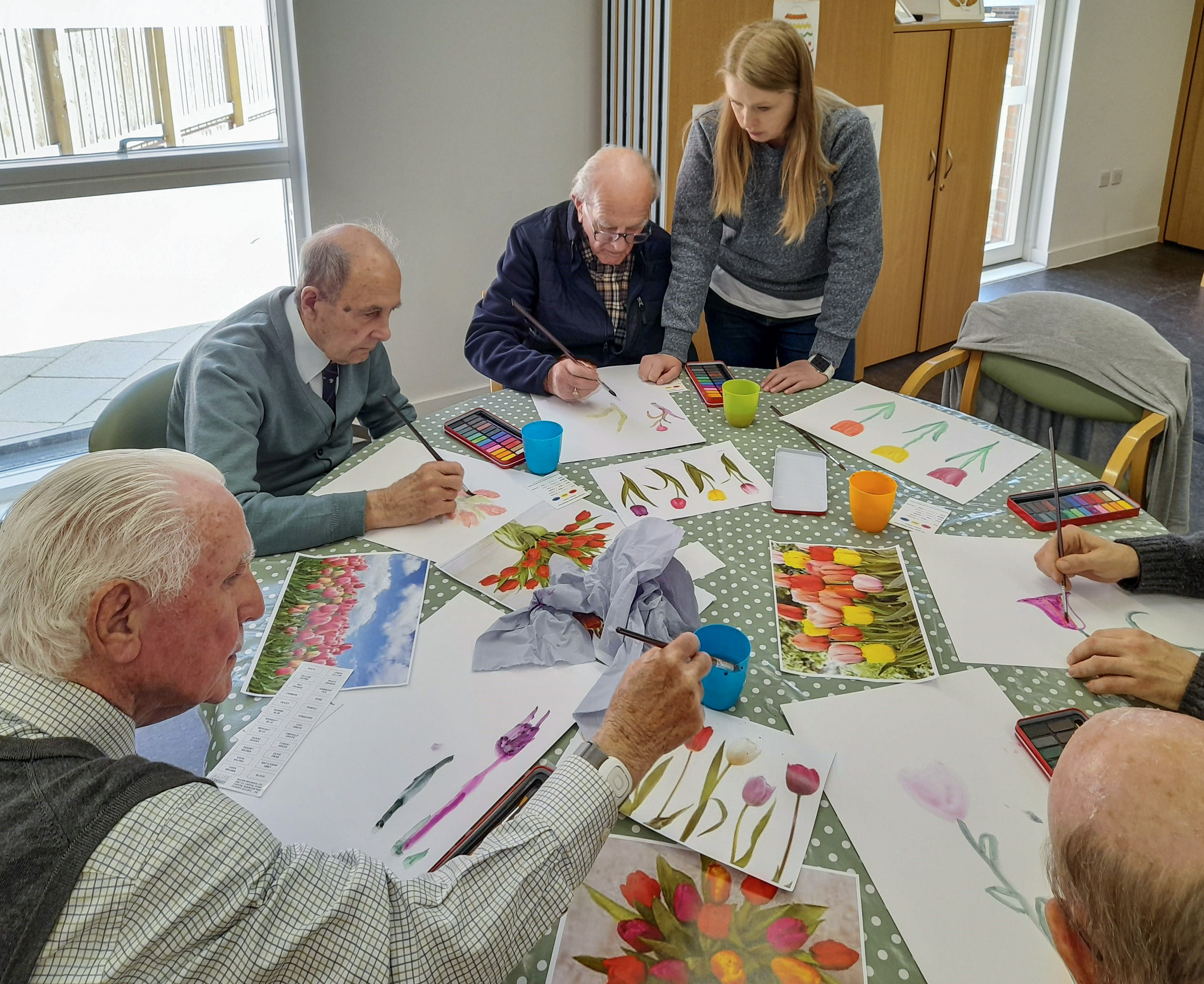 A group of older adults sitting around a table, they are painting with artist brushes, copying the colourful paintings on the table.