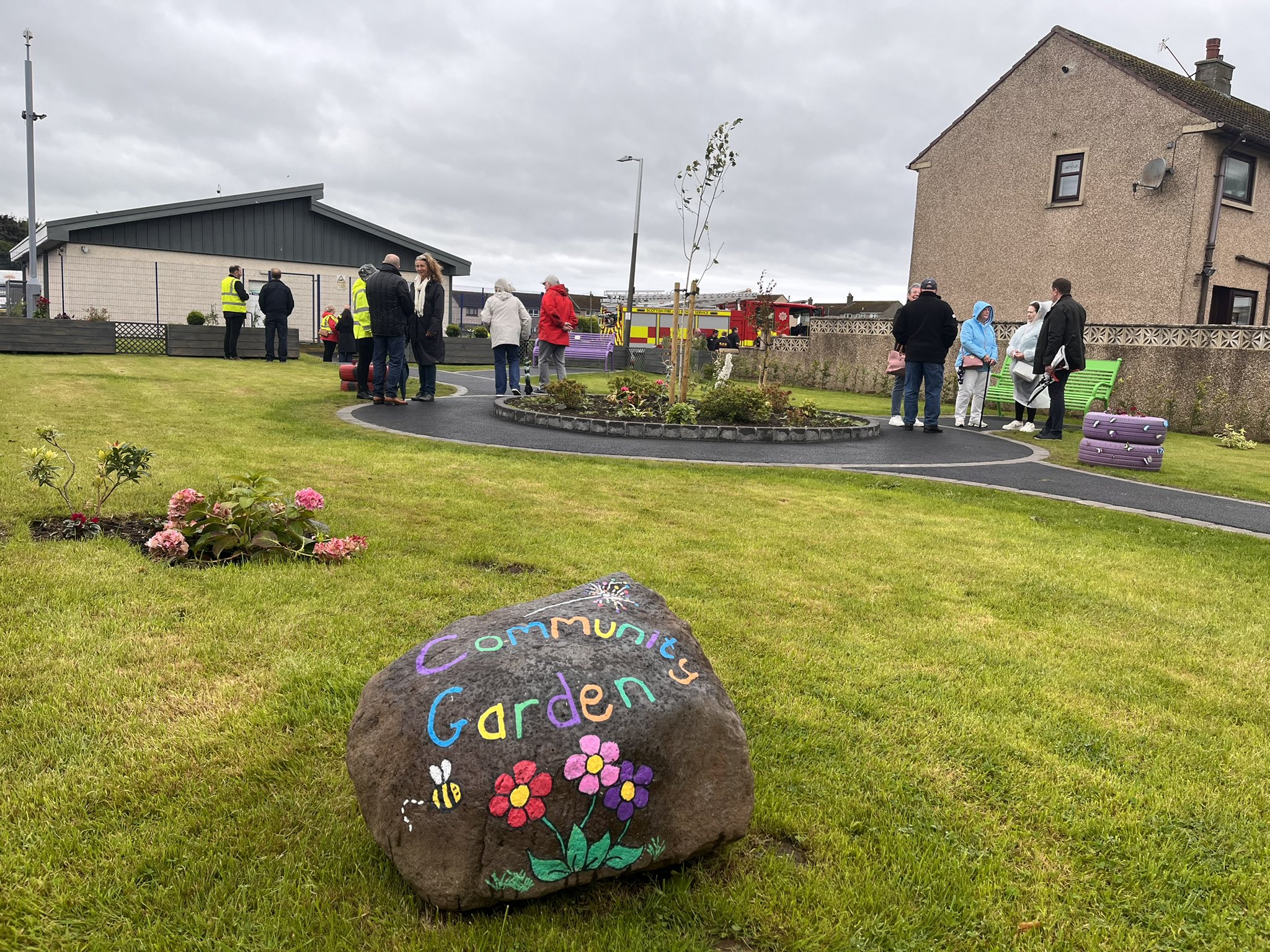 A large stone, with "Community Garden" written in bright colours, on green grass, there are large buildings and people in the background. 