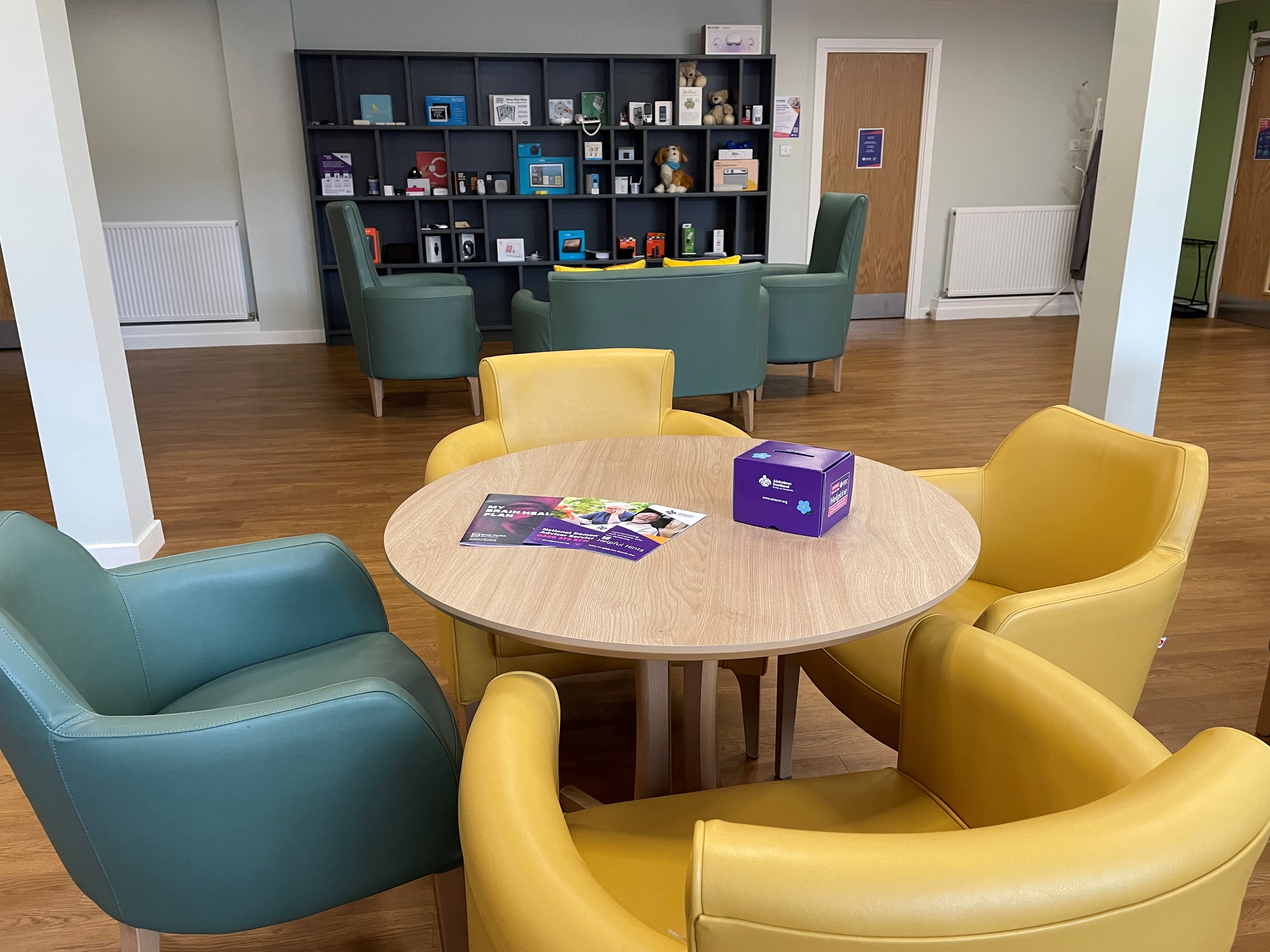 Room with wooden floors and light walls. Round table surrounded by yellow & green armchairs. Table has brochures & purple donation box. 