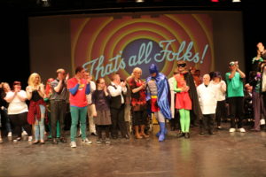 A group of people on stage in various costumes with the phrase “That’s all Folks!” projected behind them.