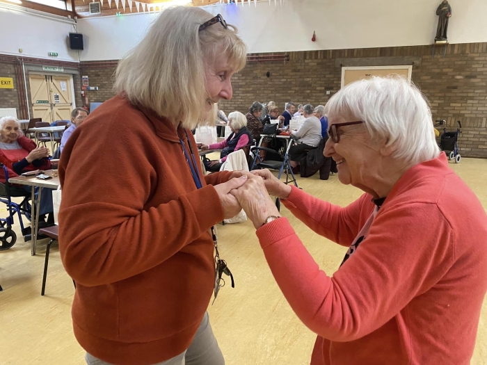 Two women dancing in a community hall with others in background