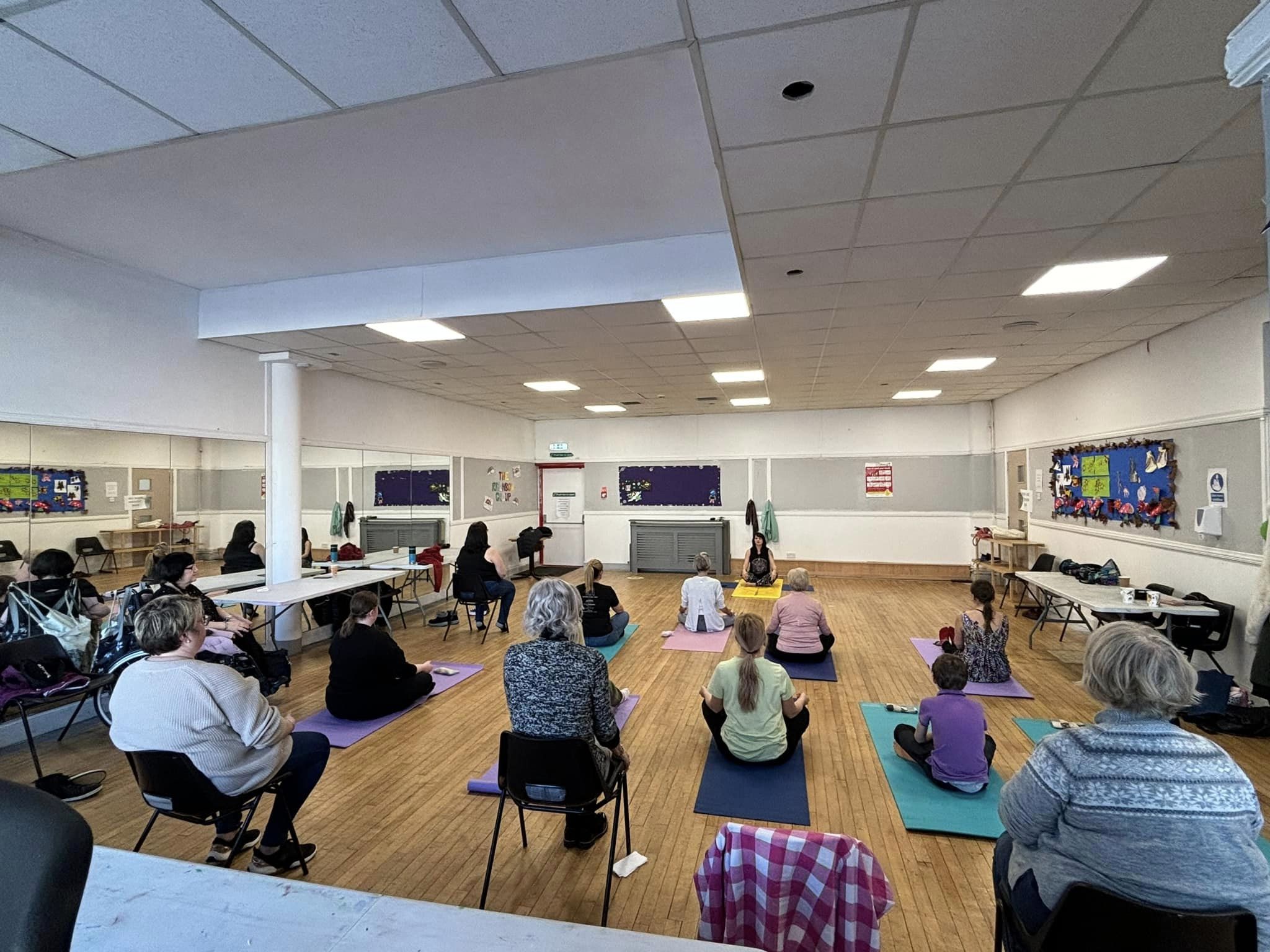 A group of people participating in a seated yoga class in a bright, spacious room with wooden floors.