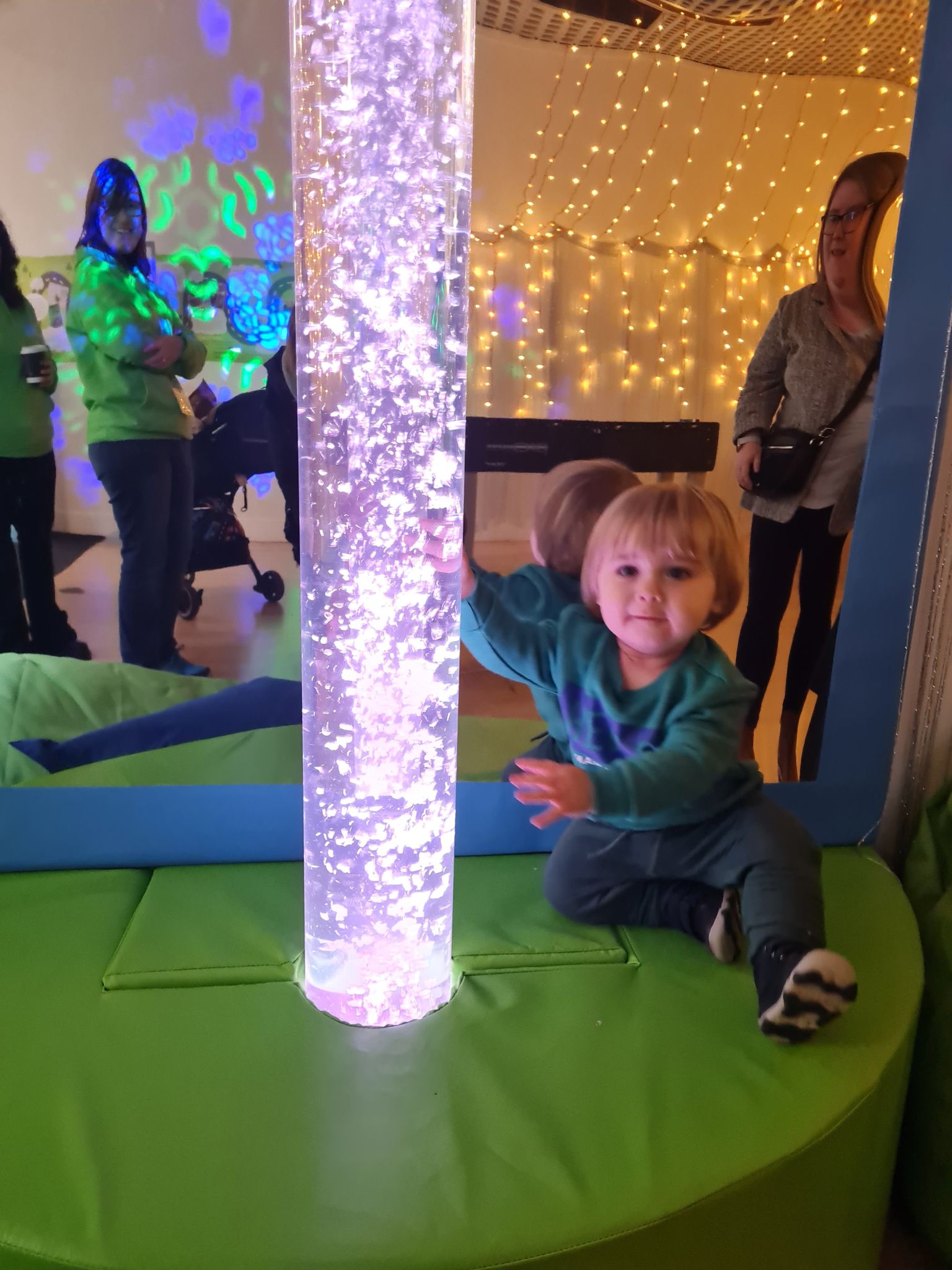 Child sitting on a raised green mat, looking at the camera, touching an illuminated pole.