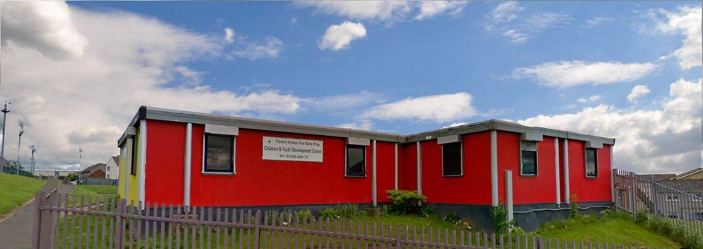 A bright red, single-story building with a sign on the front is surrounded by a grassy area and a wooden fence.