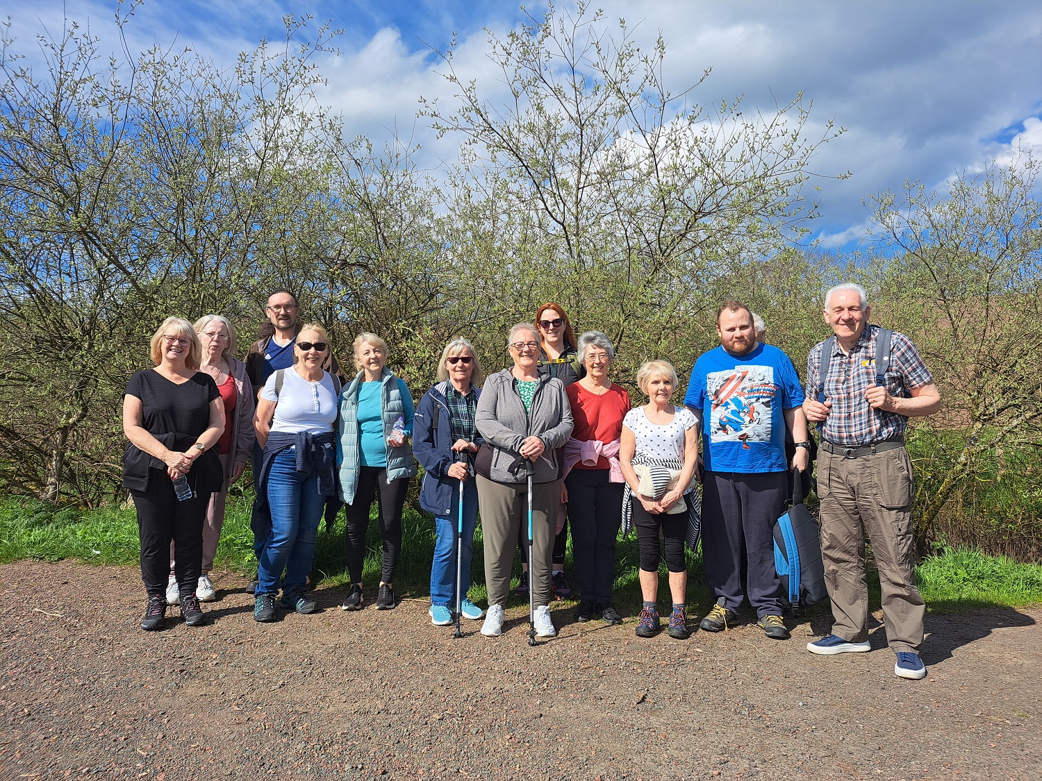 Group of walkers smiling, some younger some older. People with a walking stick. Trees in the background.