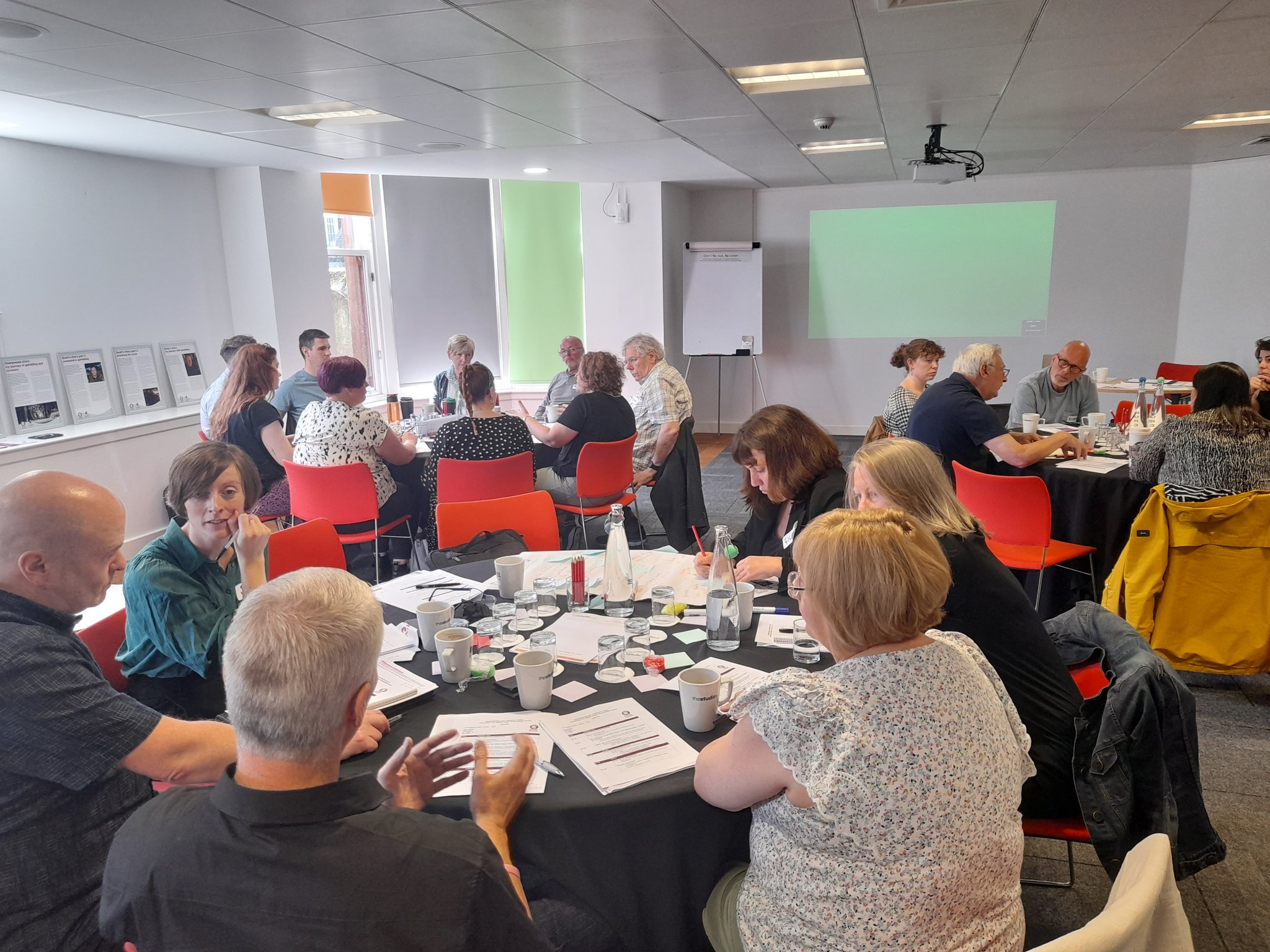 Image of roundtable event, people sitting at tables in a large room, engaged in discussion with bits of paper and pens on the tables.