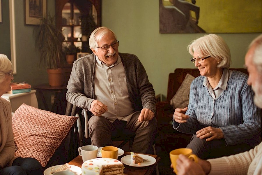 Older Man and Older Women laughing, sitting in a living room with cake and cups of tea on the table. 