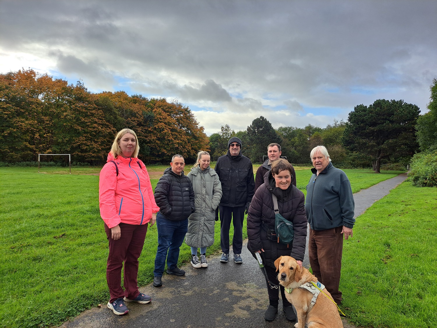 Group of walkers in a park smiling, some younger some older. Person with a guide dog.