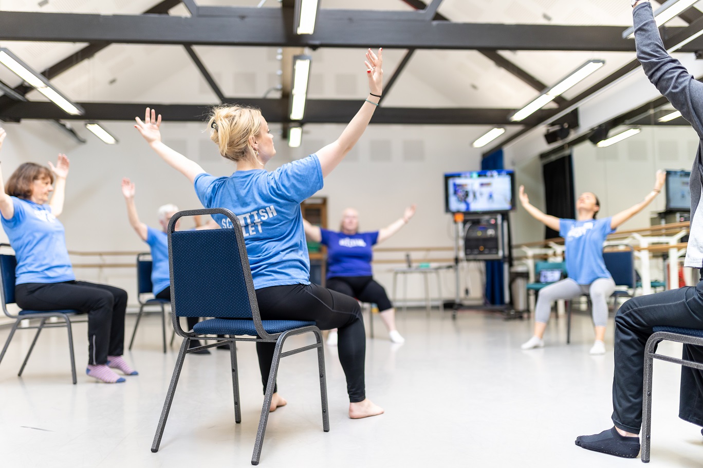 Seated dancers wearing light blue Scottish Ballet t-shirts, sit on chairs in a circle with their arms outstretched