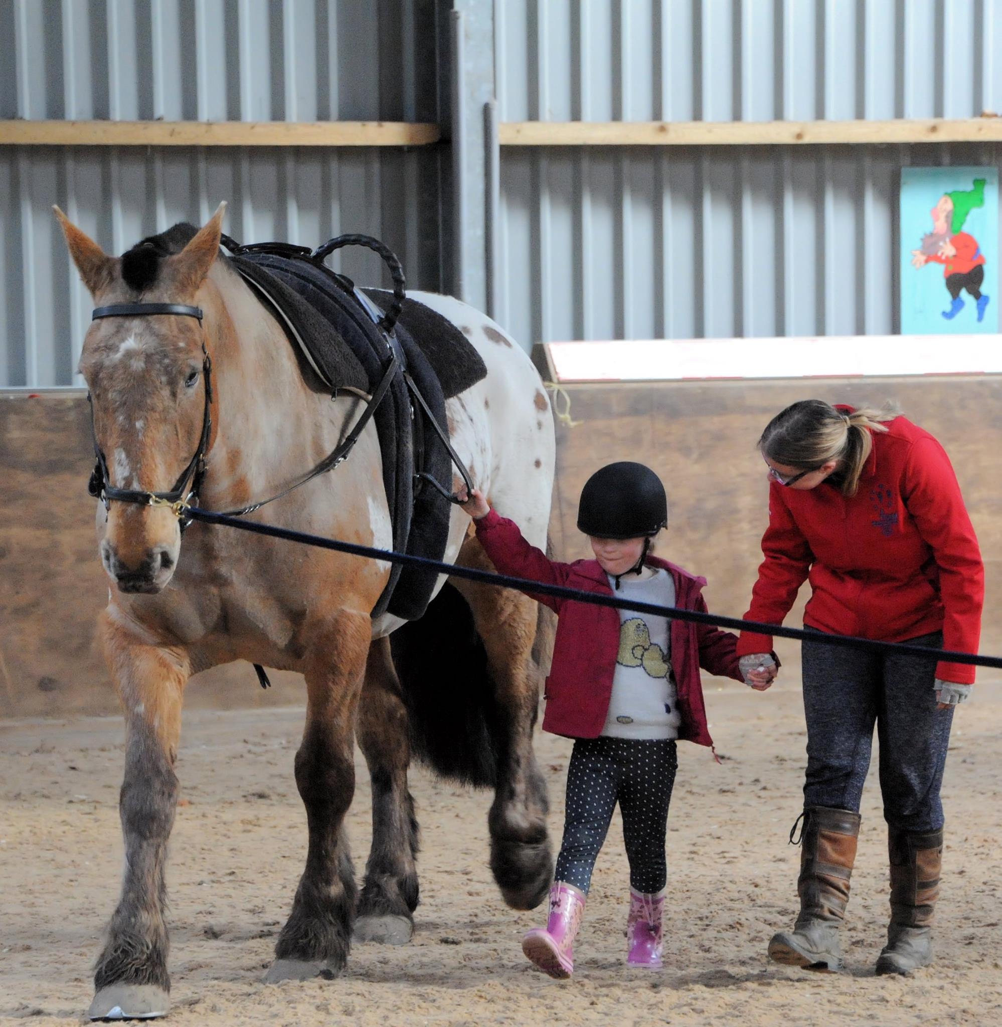 A small child and adult walking next to a horse in a large barn.