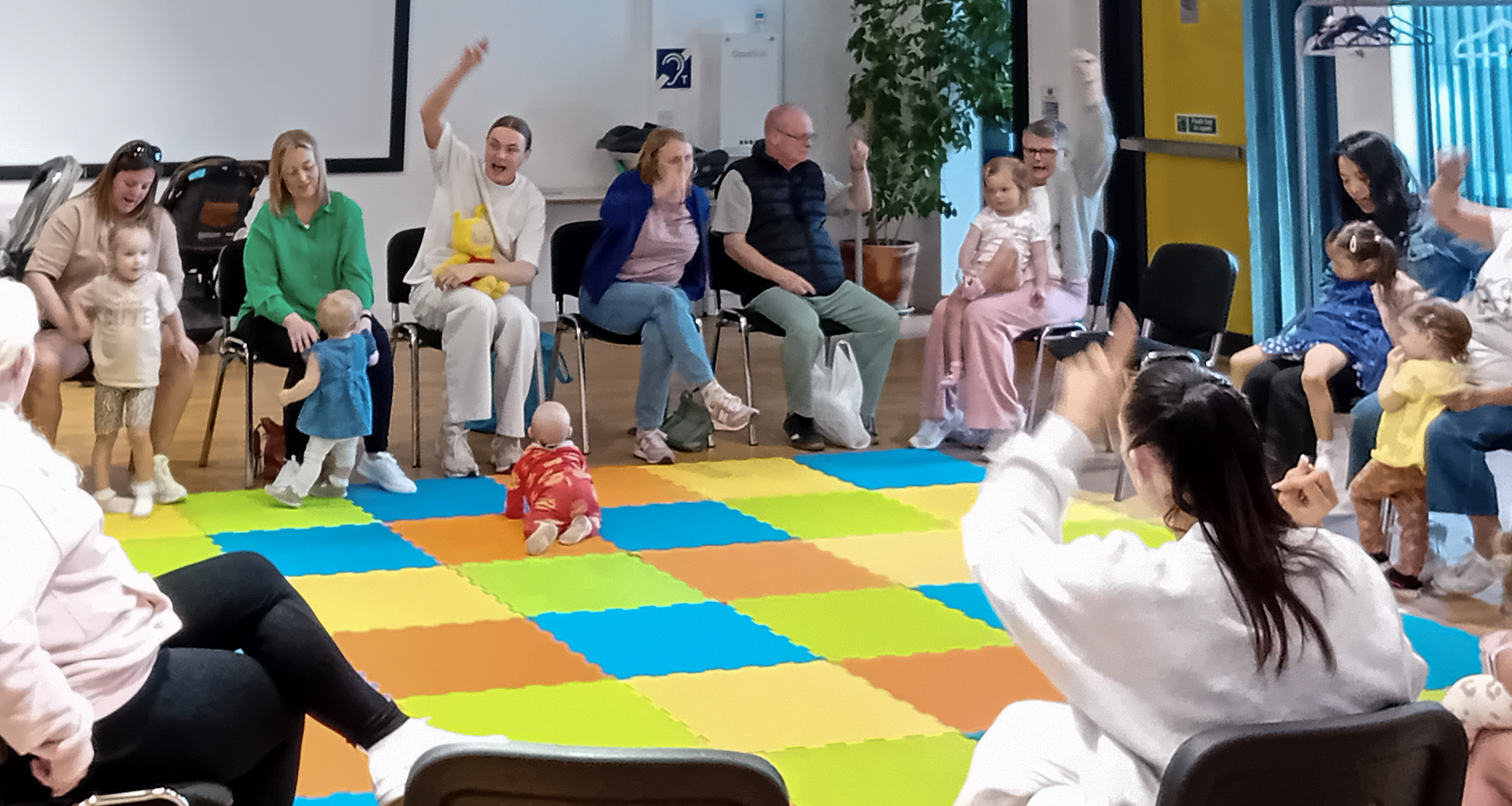 Large room with multicoloured flooring. Parents and guardians are seated in a circle watching children play.