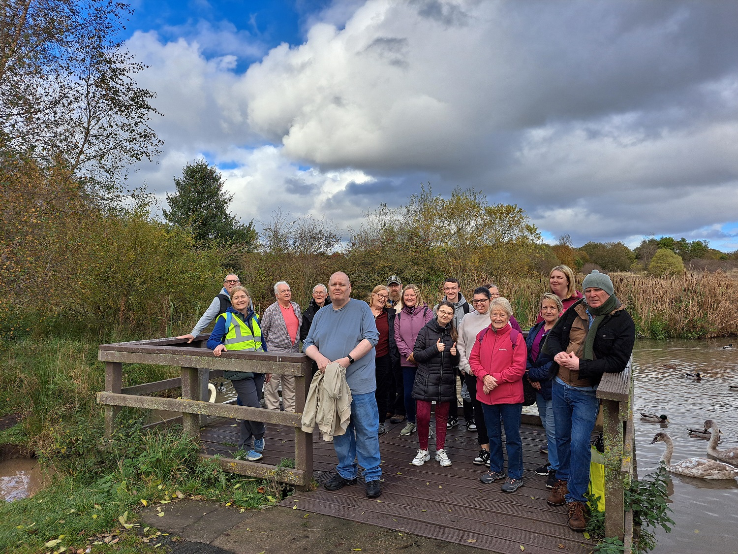 Group of walkers in a park smiling, some younger, some older. Pond with ducks and swans in the background.