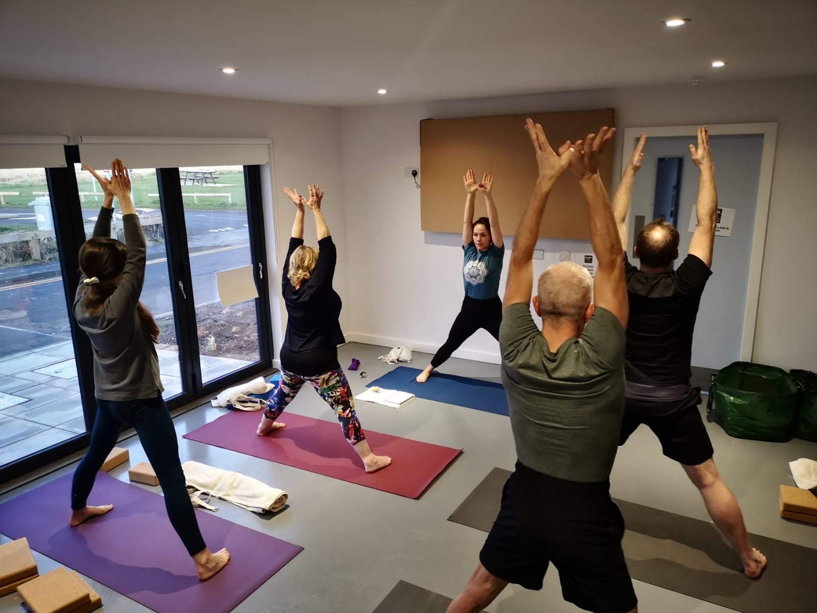 Five people standing on colourful mats with their legs wide and arms in the air, there's a large window letting in lots of light to the room