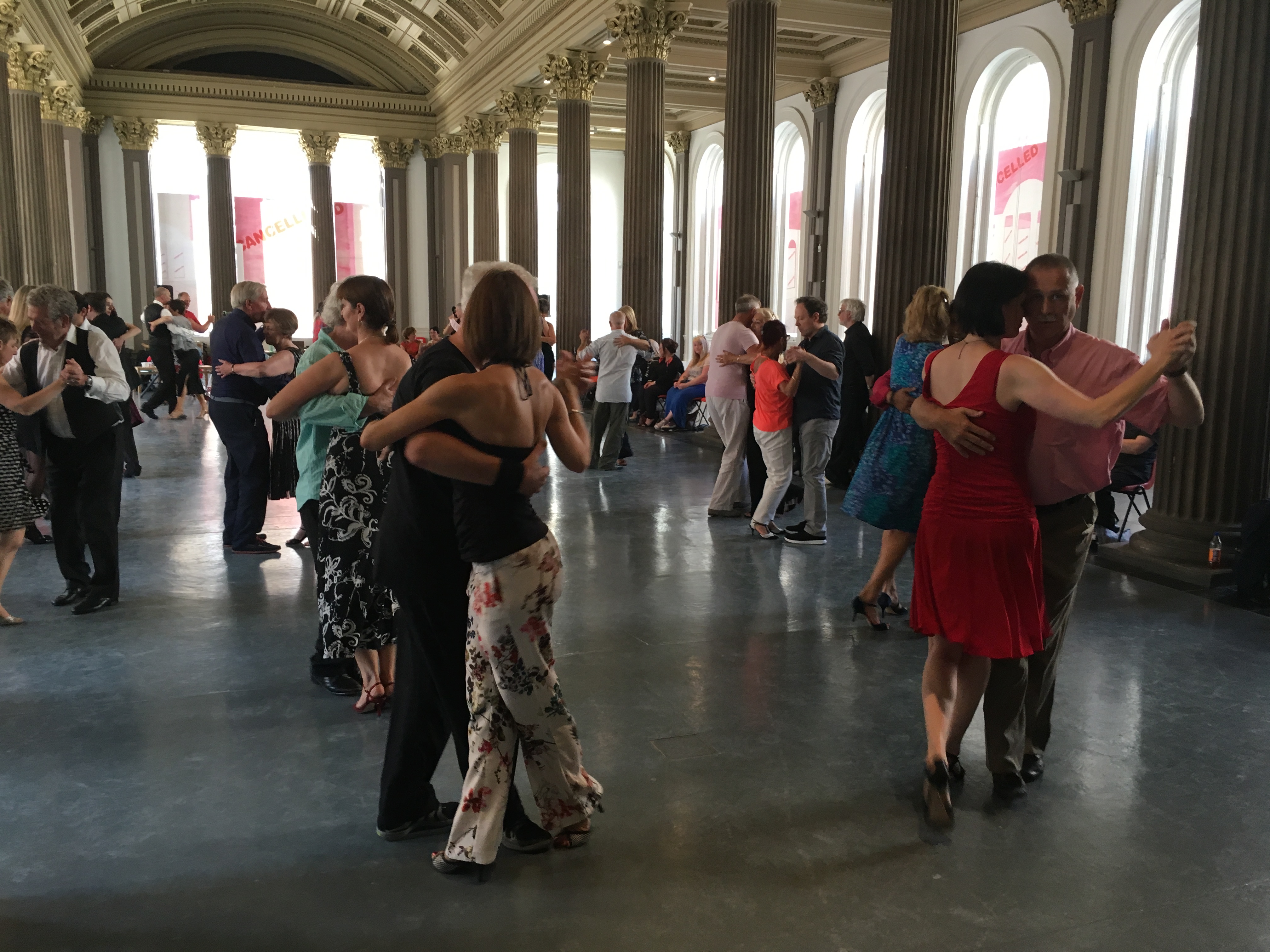 People dancing the tango in a large bright room with lots of windows with stone pillars.