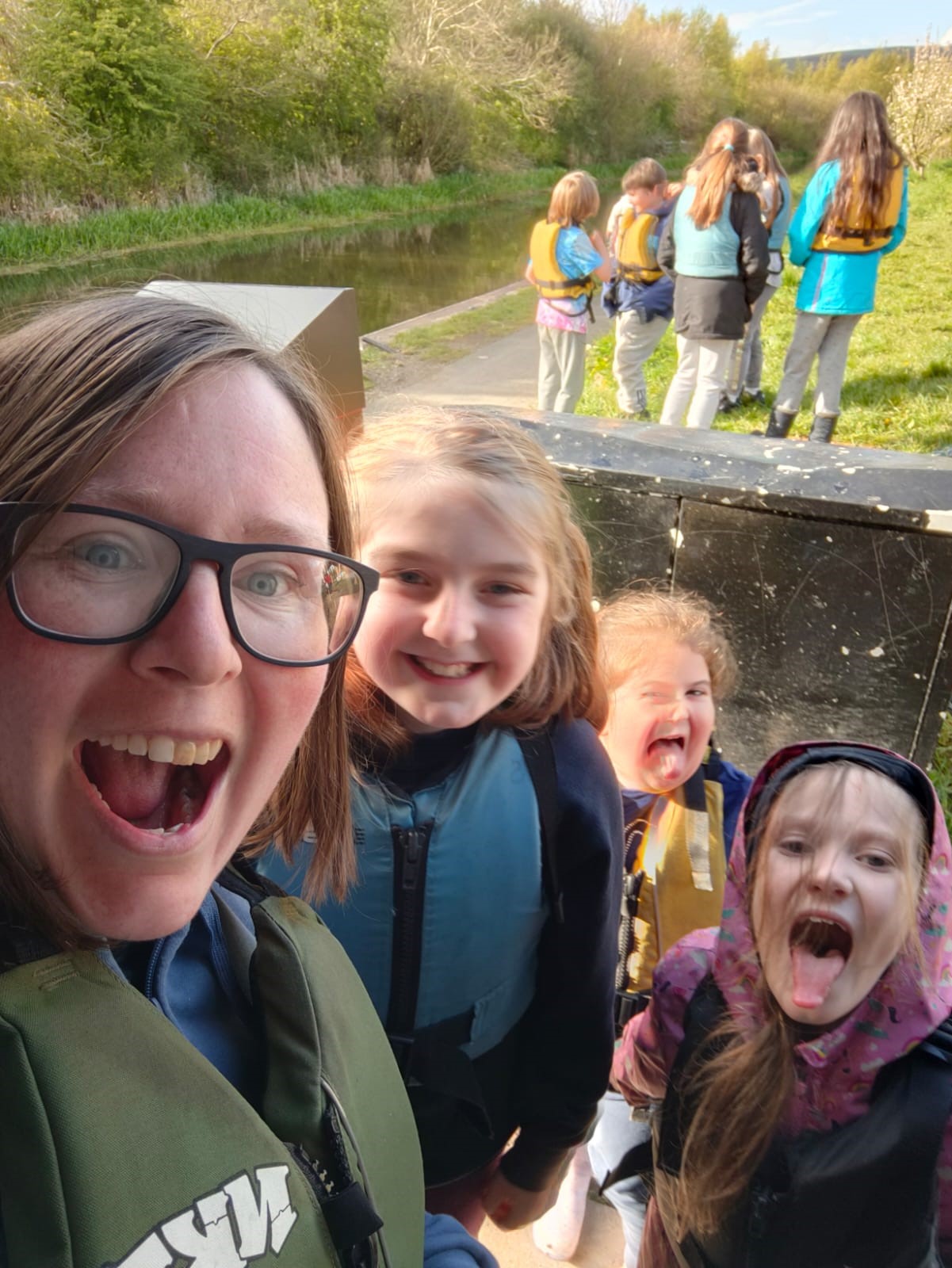 A group selfie of a smiling adult & 3 excited children in life jackets by a canal, more kids in life jackets gathered in the background. 