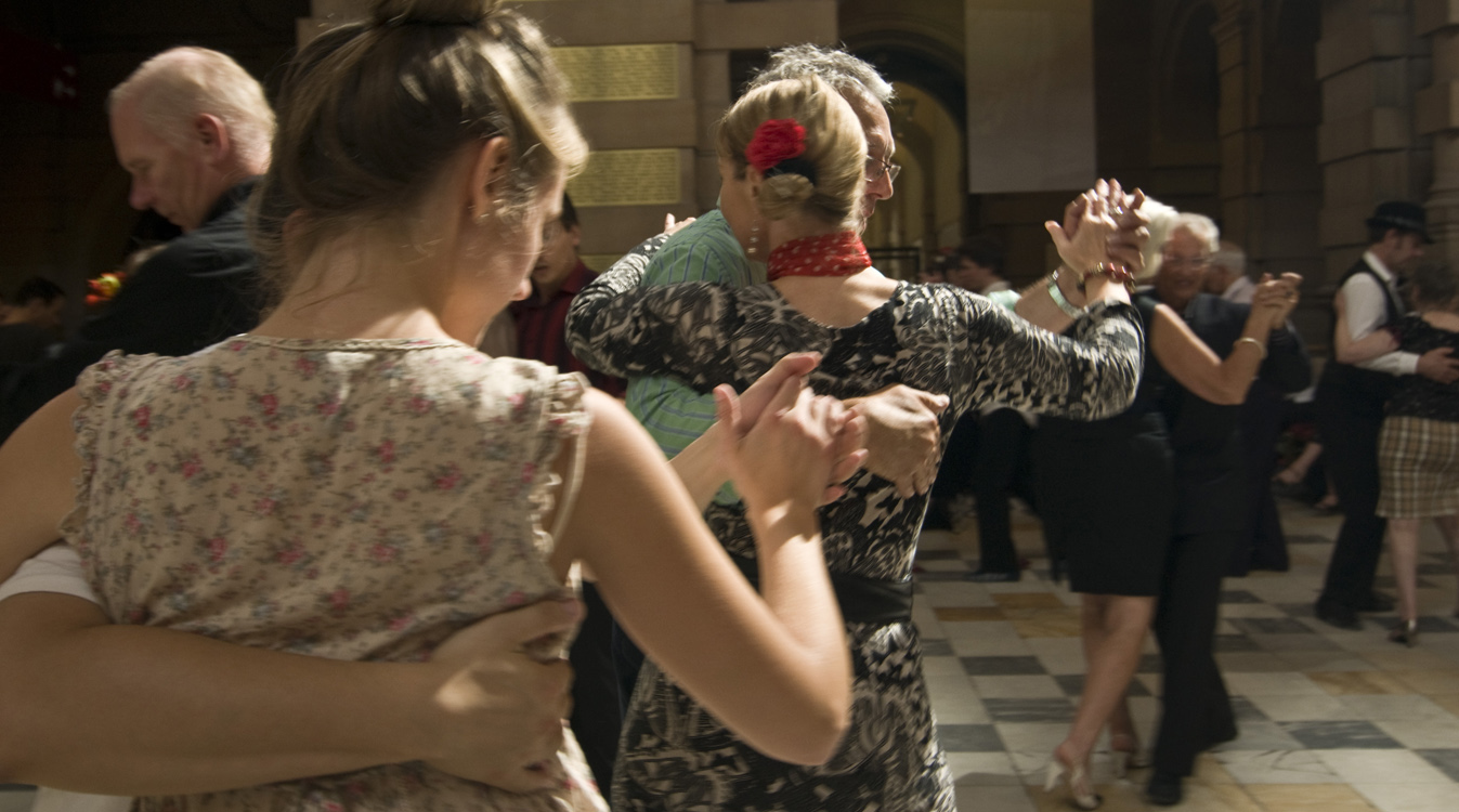 Close up of people dancing the tango in a large room with a checked floor.