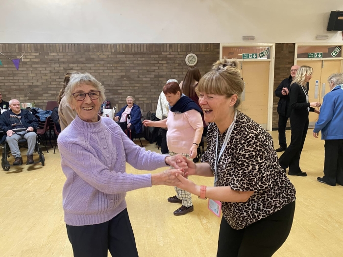 Two women dancing in a community hall with others in background