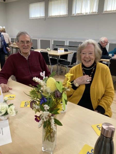 Two people sitting at a table with a vase of yellow and green flowers, they are both smiling at the camera.