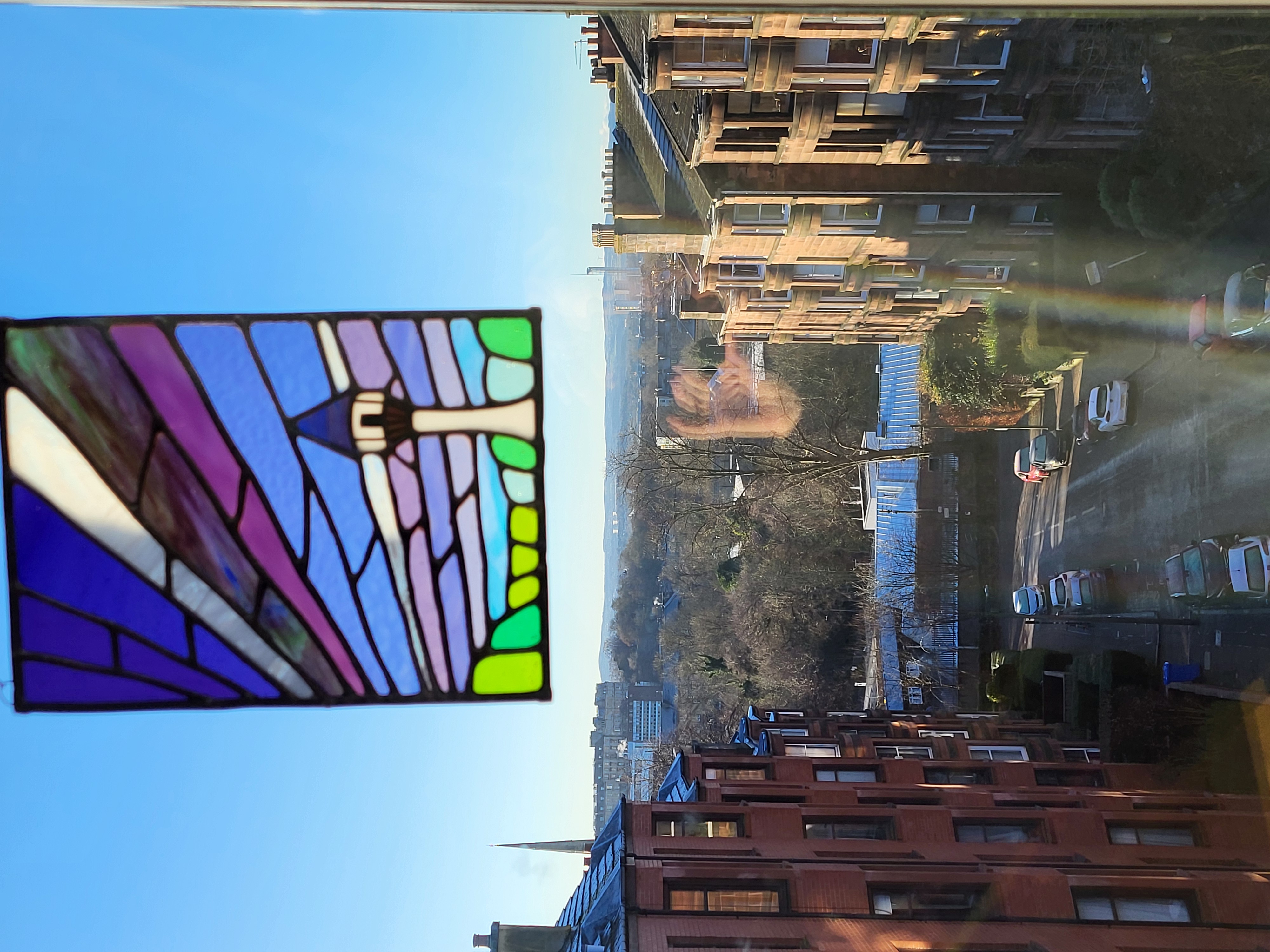 View looking into a street with red stone tenements and park at the end. A colourful stained glass window ornament at the top of the window