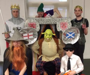 A group of people in medieval costumes, holding shields and wearing crowns, with their faces obscured.