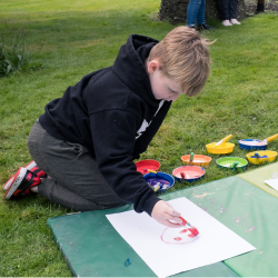 Child kneeling on the grass in black hooded top paints a red smiling face on to paper with a brush. different coloured paints in pots behind