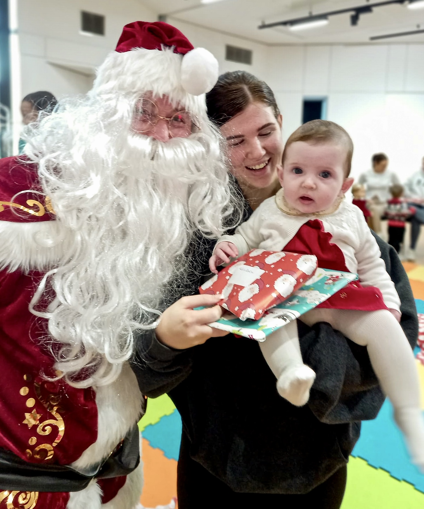 Santa Claus smiling at the camera standing next to an adult holding a small child in a red dress, who looking into the camera.