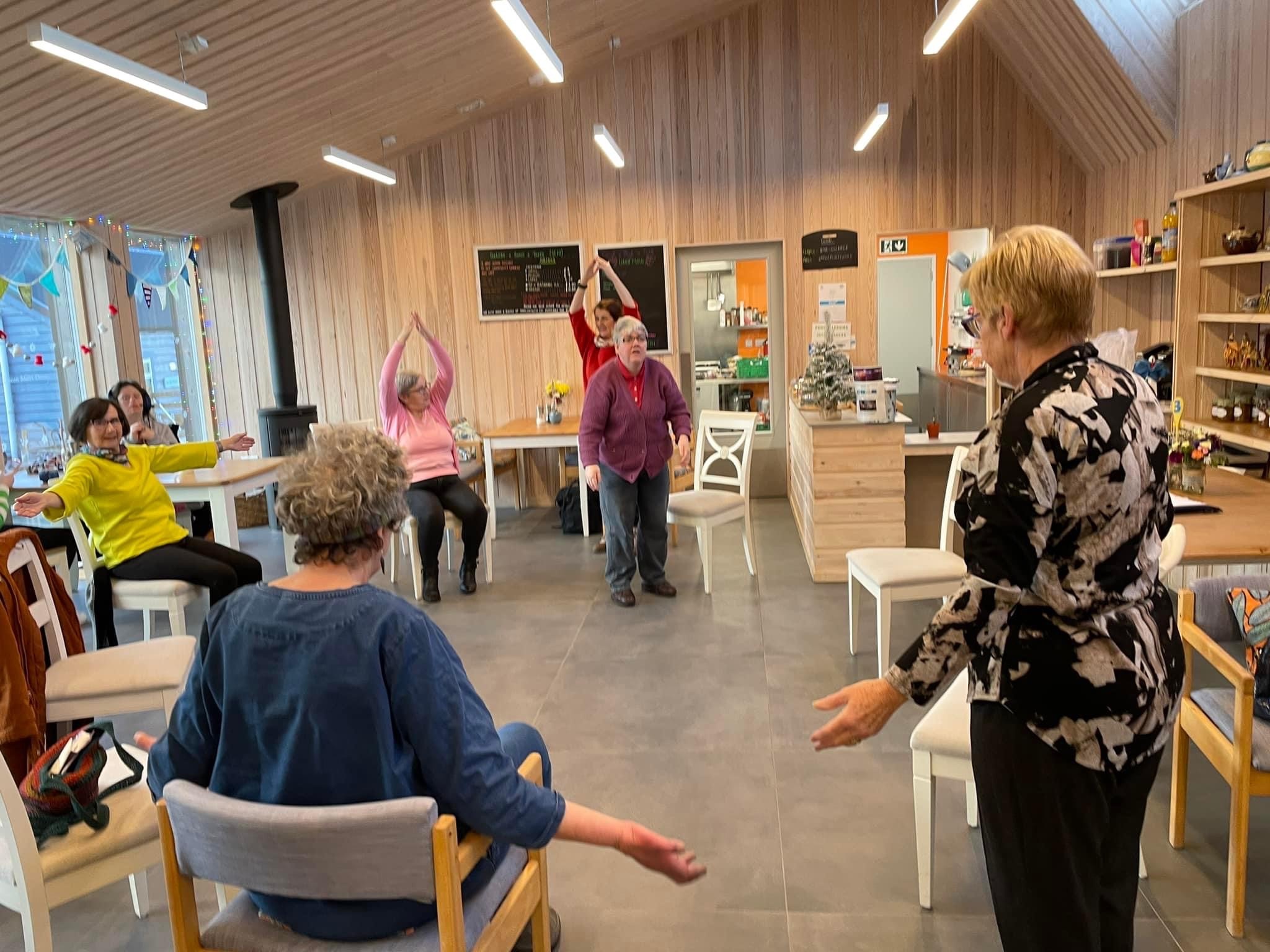 A large bright room with peoople sitting and standing in a circle moving their arms.