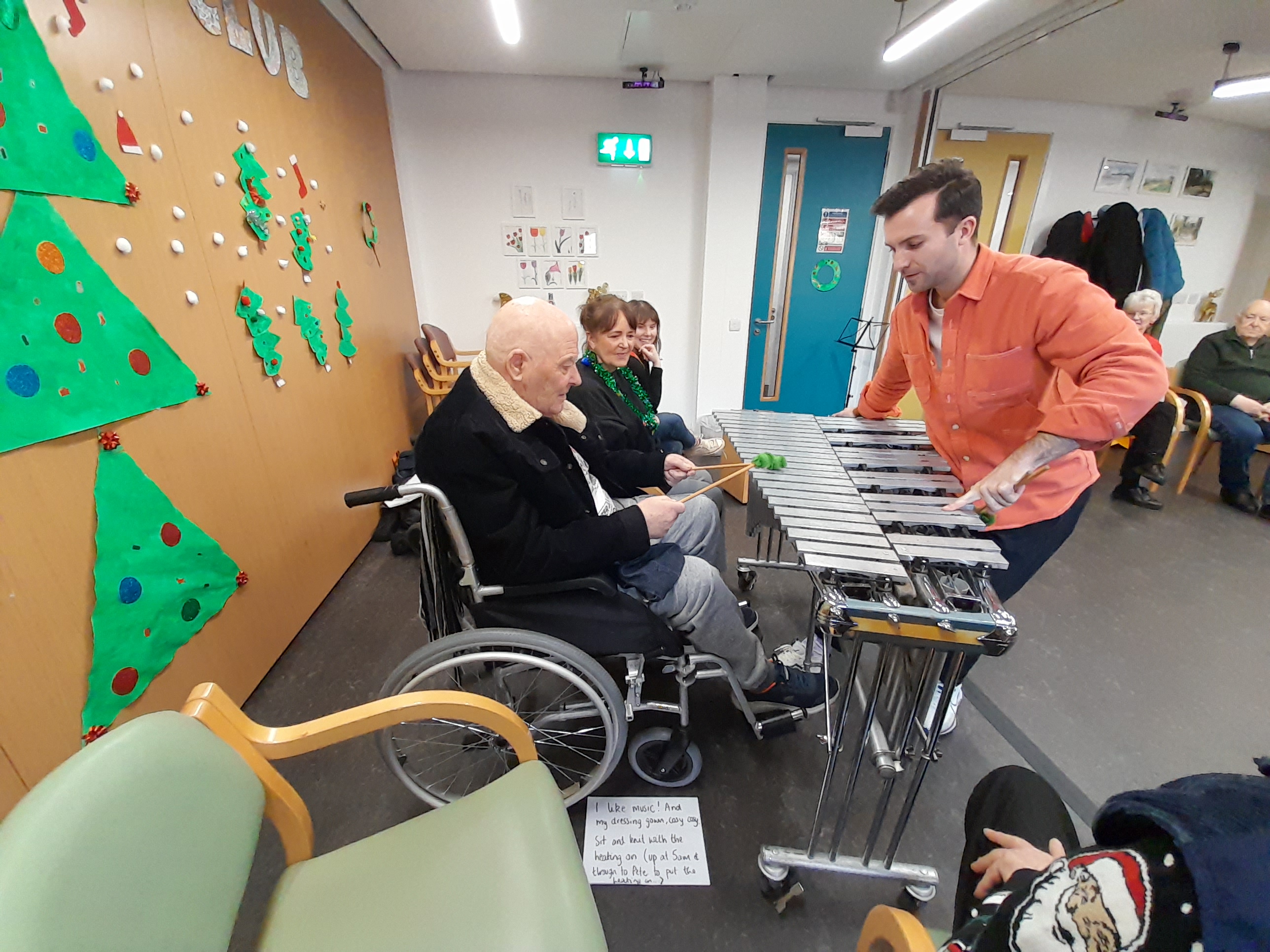 Older adult in a wheelchair learning to play the Xylophone by a younger adult with an organg shirt on. The room is bright.