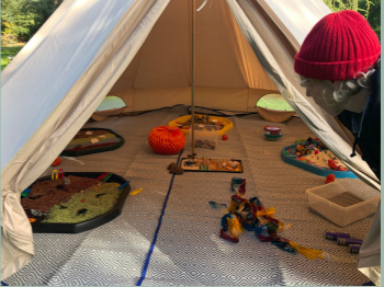 A Bell tent with tuff trays containing sensory items like coloured rice and toys, on the floor of the tent is a mat with toys.