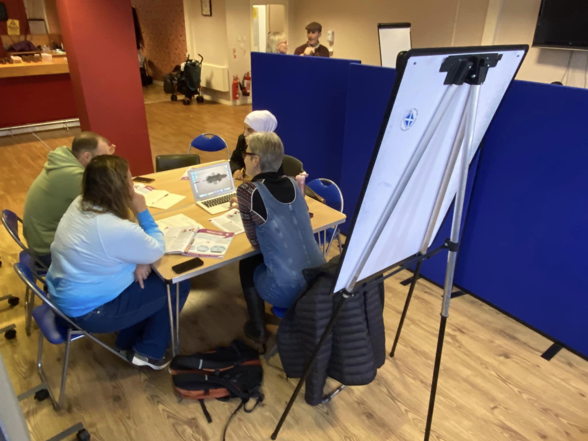 People sitting at a table with a flipchart at the top of the table. The people are in a large room with red pillars.