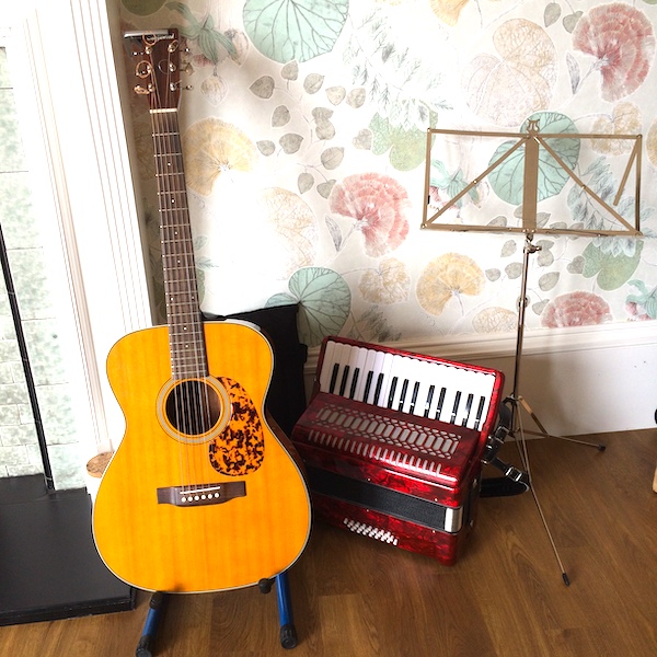 A wooden guitar  on a stand and an accordion sitting next to the stand on the floor.