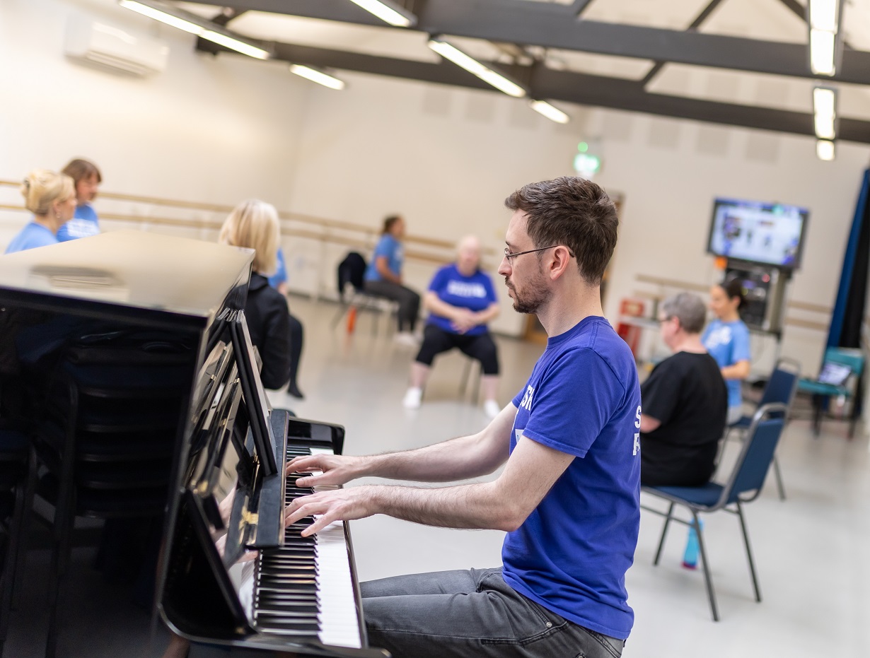 A pianist plays in the foreground, in the background are seated people taking part in a movement class