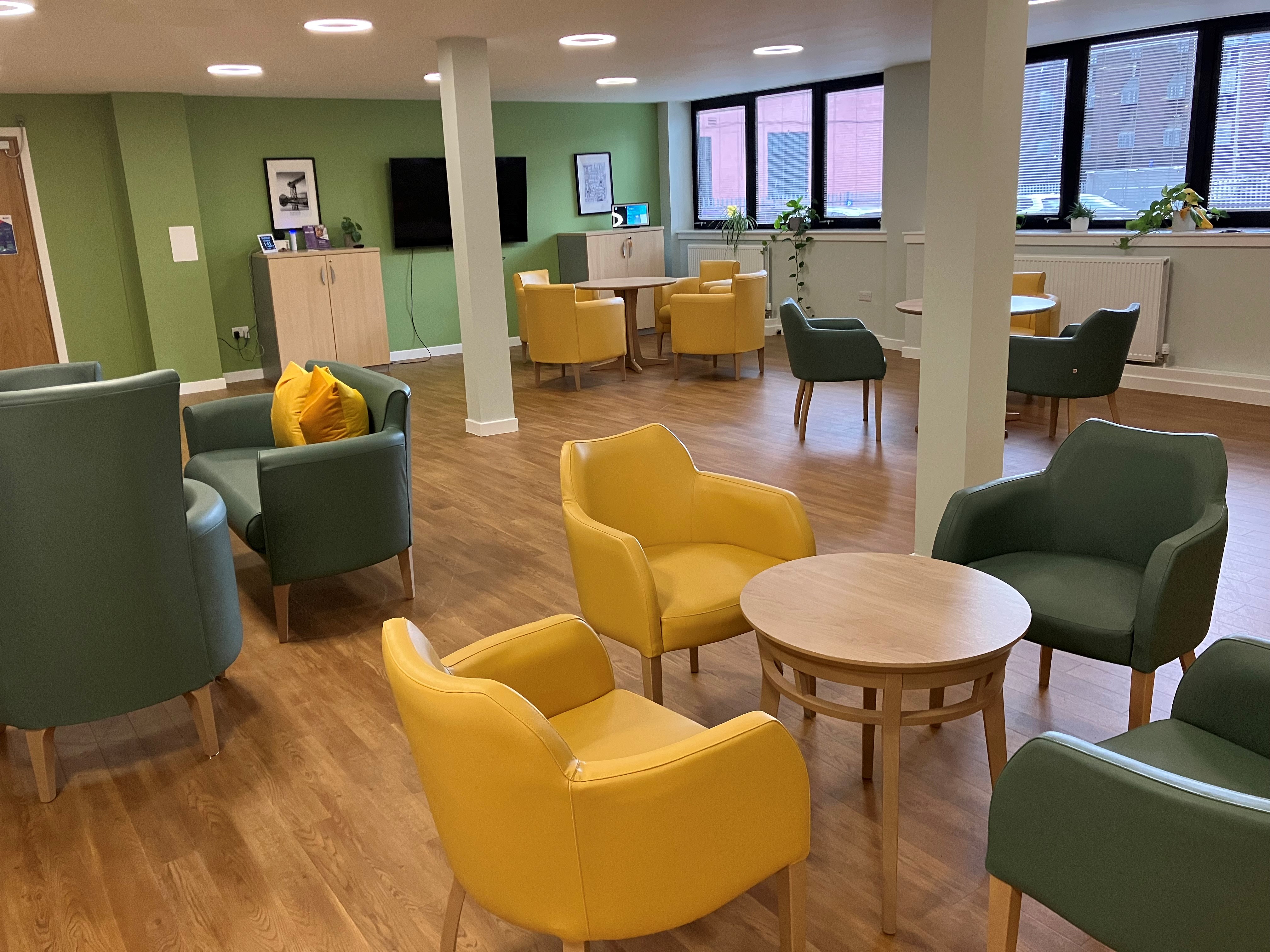 A cosy, well-lit room with wooden floors and light green walls. There are green and yellow armchairs around small wooden tables.