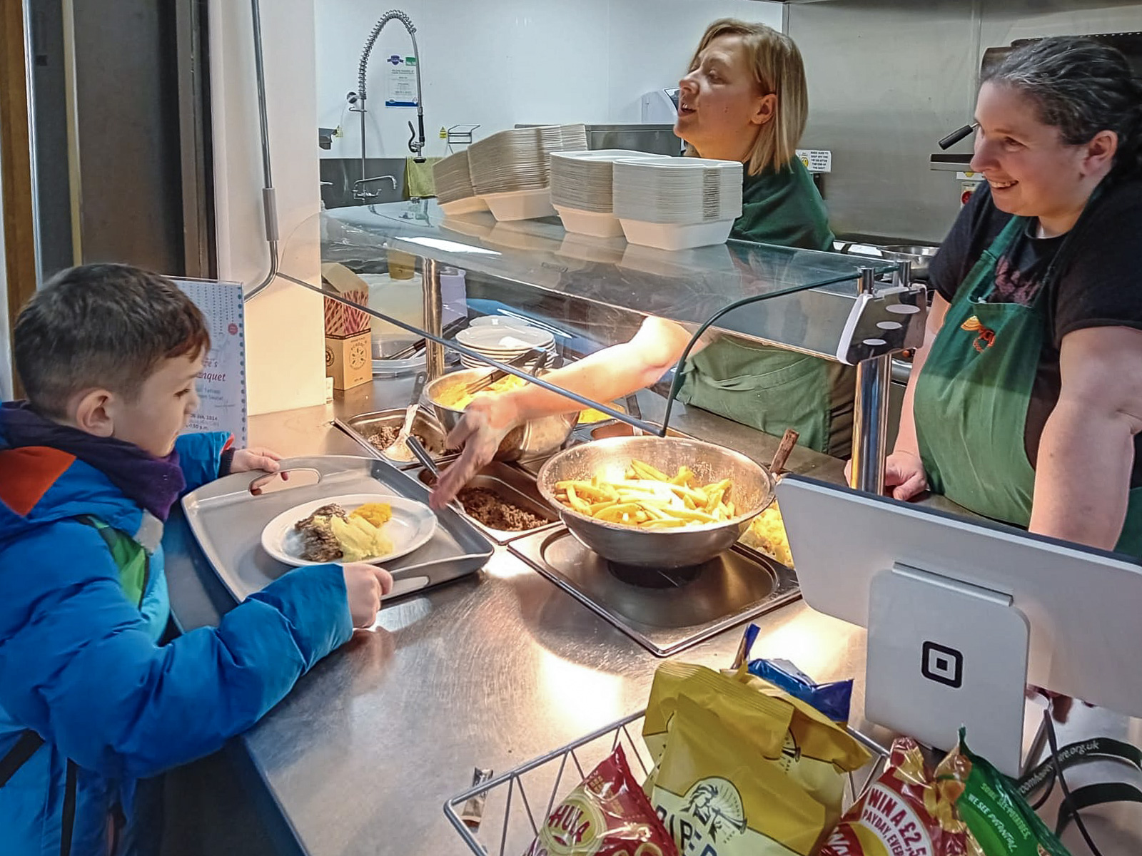 A child with a tray is being served food by two smiling adults in aprons behind a counter, with dishes and snacks displayed nearby.