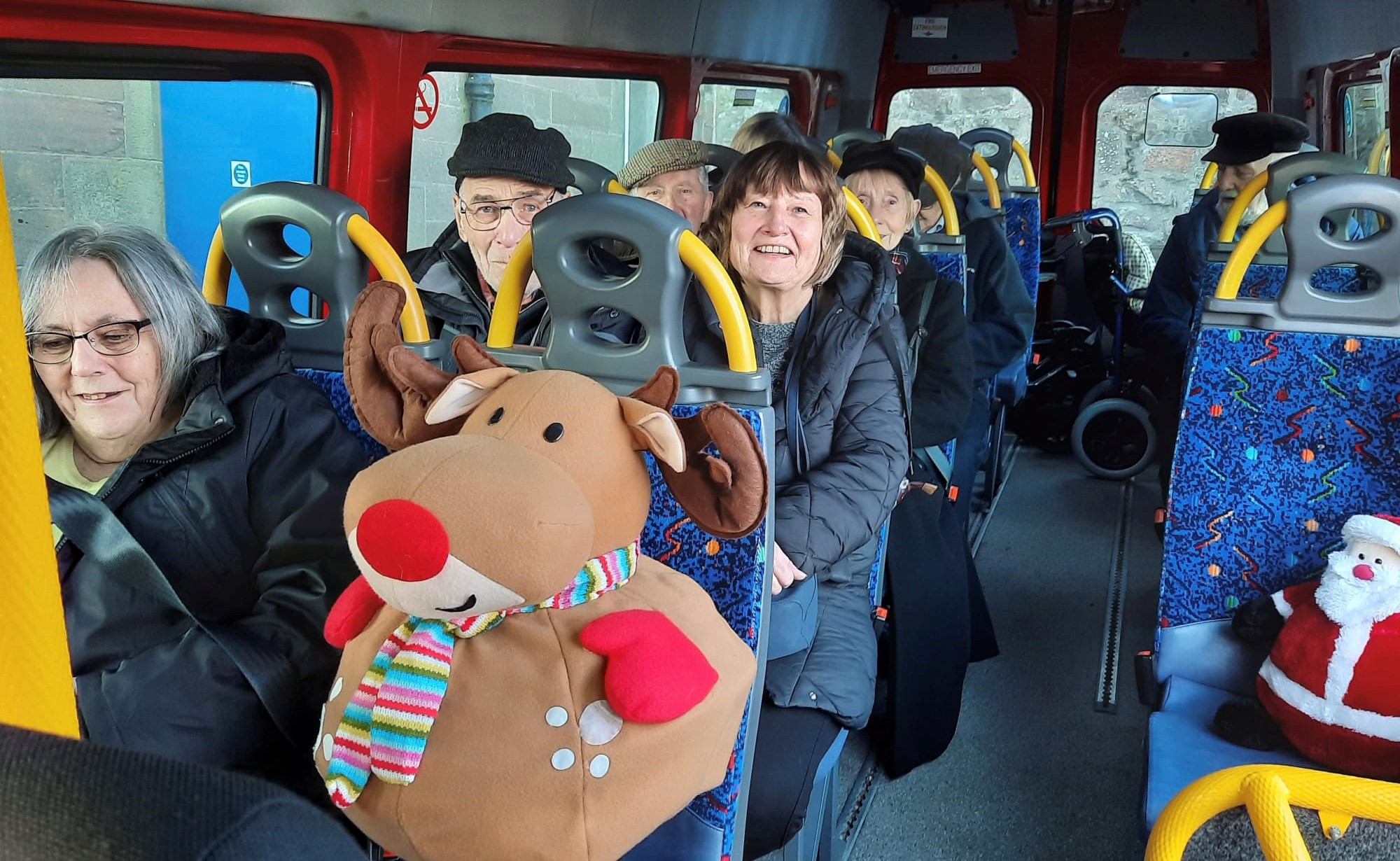 Group members and a large stuffed toy sitting on a bus that has blue and yellow seats.