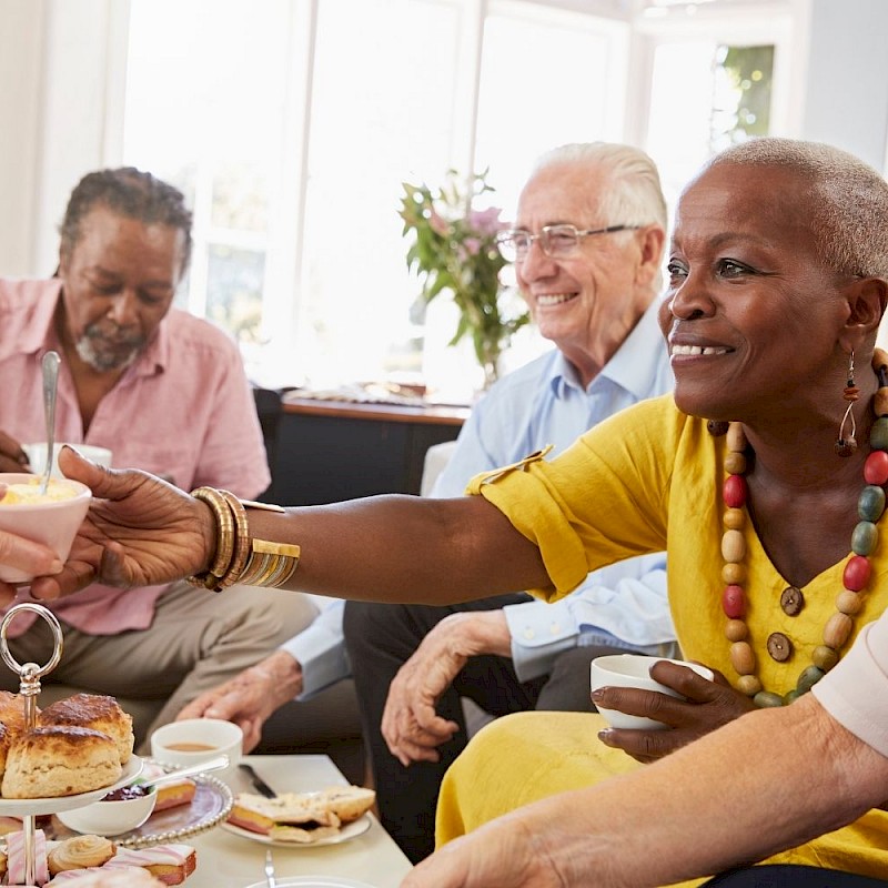 Older lady reaching across a coffee table for a cup of coffee. There are 2 older men in the background smiling.