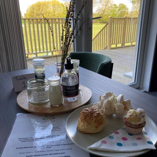 Cafe table with a menu, condiments and plate with three cakes and a spotty napkin.  Window looking out to decking.